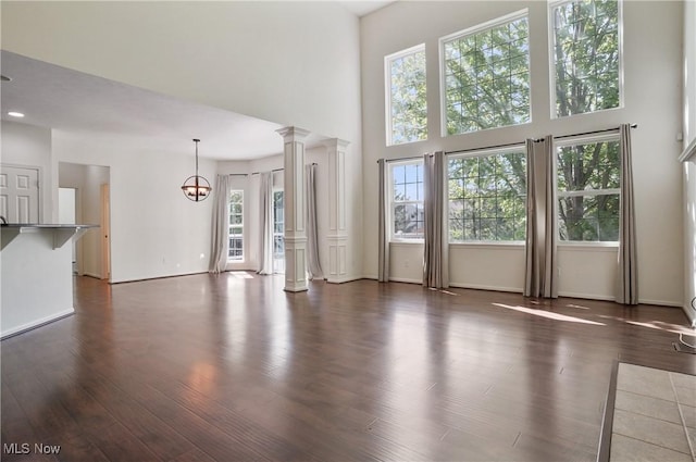 unfurnished living room featuring decorative columns, dark wood finished floors, a towering ceiling, baseboards, and a chandelier