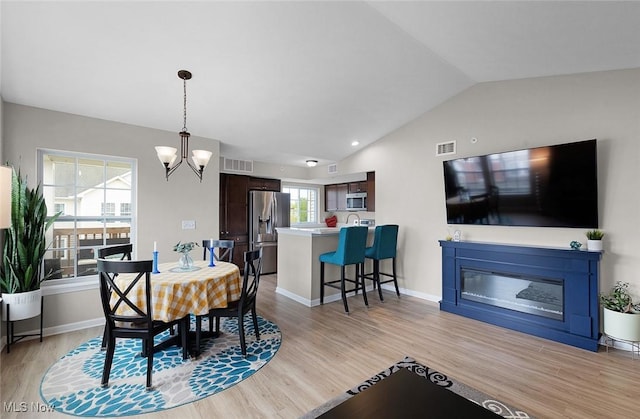 dining space featuring vaulted ceiling, light wood-style flooring, a chandelier, a glass covered fireplace, and baseboards