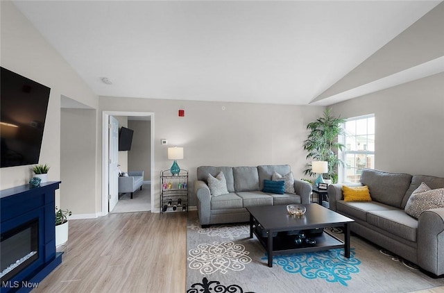 living area featuring vaulted ceiling, light wood-style flooring, a glass covered fireplace, and baseboards