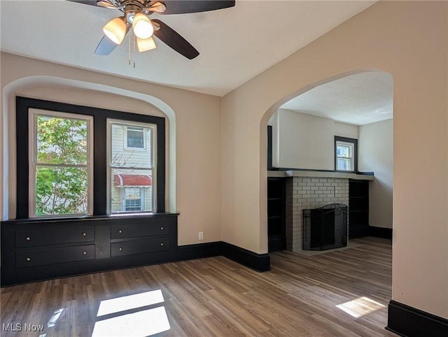 empty room featuring wood finished floors, a ceiling fan, baseboards, a fireplace, and arched walkways