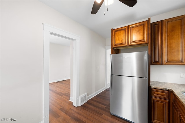 kitchen with freestanding refrigerator, ceiling fan, dark wood-style flooring, light stone countertops, and brown cabinets