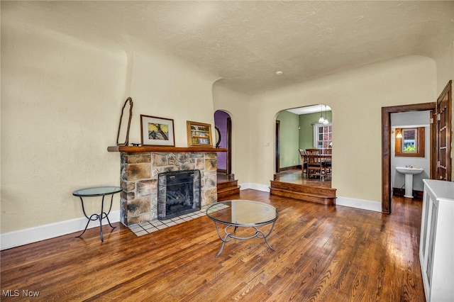 living room featuring arched walkways, wood finished floors, a stone fireplace, baseboards, and a textured ceiling