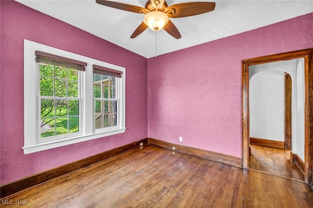empty room featuring wood finished floors, baseboards, a textured wall, ceiling fan, and arched walkways