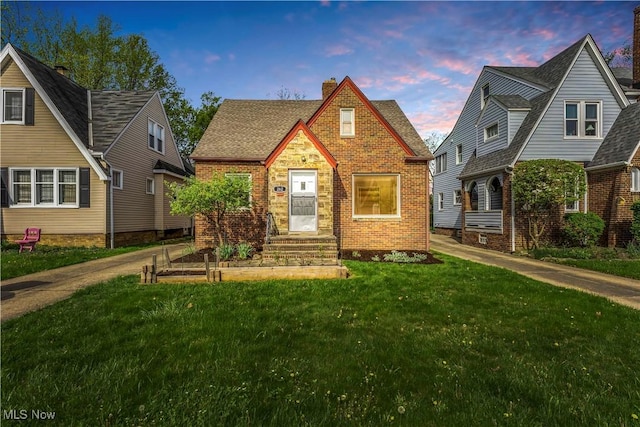 tudor house featuring a chimney, brick siding, a yard, and roof with shingles