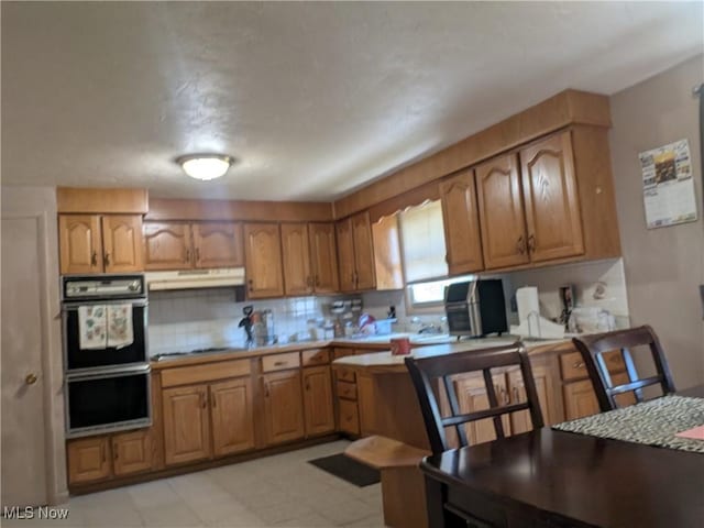 4222 Kenyon Ct, Lorain, OH 44053 - kitchen featuring brown cabinets, light countertops, backsplash, under cabinet range hood, and multiple ovens