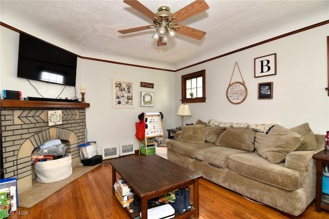 living room with a ceiling fan, wood finished floors, ornamental molding, a brick fireplace, and a textured ceiling