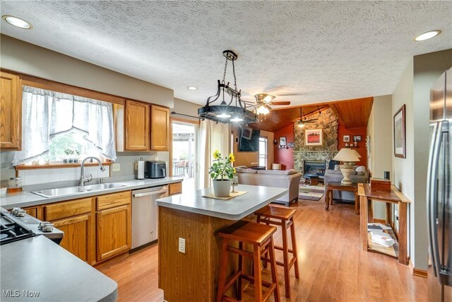 kitchen featuring stainless steel appliances, a stone fireplace, a sink, a ceiling fan, and a kitchen island