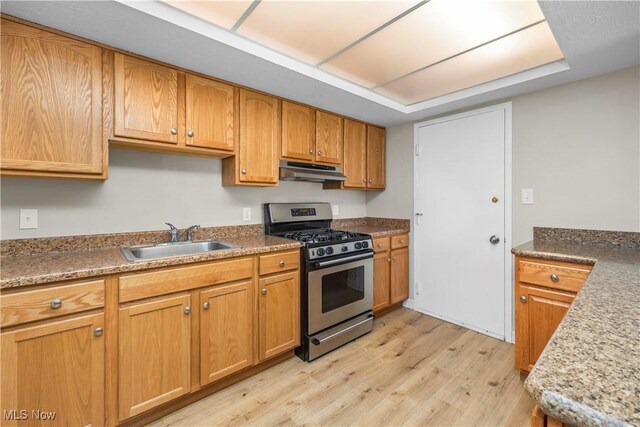kitchen featuring stainless steel range with gas cooktop, a sink, under cabinet range hood, light wood-style floors, and brown cabinetry