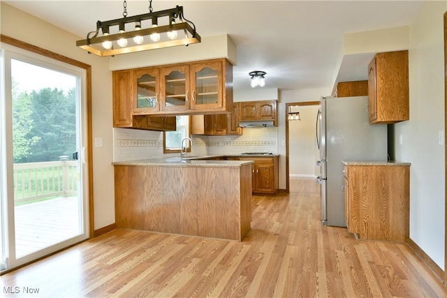 kitchen with brown cabinetry, freestanding refrigerator, under cabinet range hood, a peninsula, and tasteful backsplash