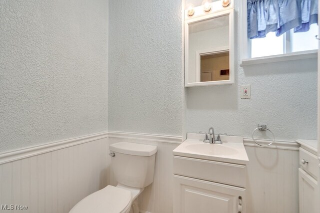 half bath with a wainscoted wall, a textured wall, vanity, and toilet