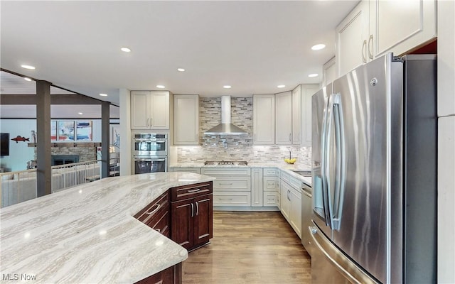 kitchen featuring stainless steel appliances, wall chimney exhaust hood, light stone countertops, decorative backsplash, and wood finished floors