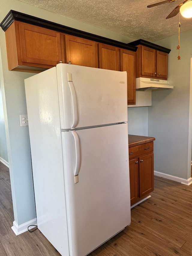 kitchen with a textured ceiling, under cabinet range hood, freestanding refrigerator, brown cabinets, and dark wood finished floors