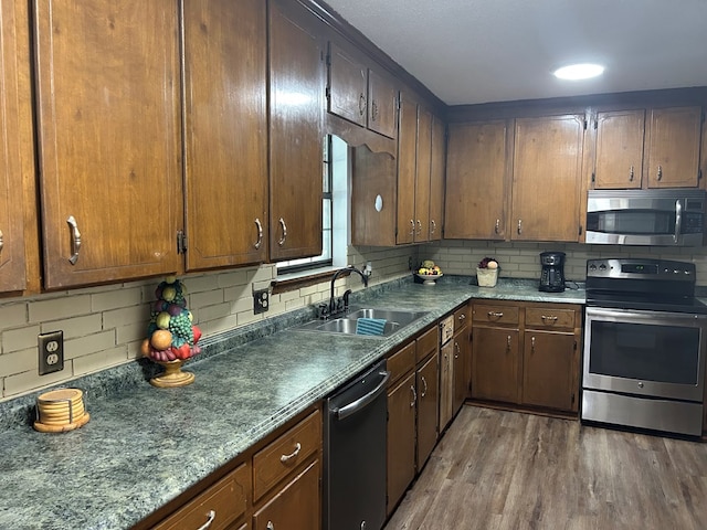 kitchen featuring stainless steel appliances, wood finished floors, a sink, backsplash, and dark countertops