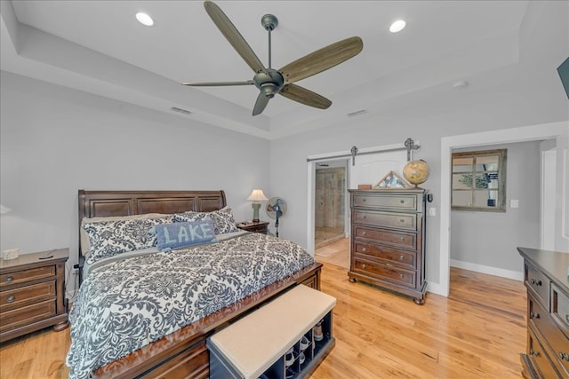 bedroom with ensuite bath, ceiling fan, a tray ceiling, light hardwood / wood-style floors, and a barn door