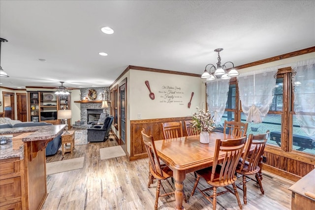 dining room featuring ceiling fan with notable chandelier, a stone fireplace, wood walls, ornamental molding, and light wood-type flooring