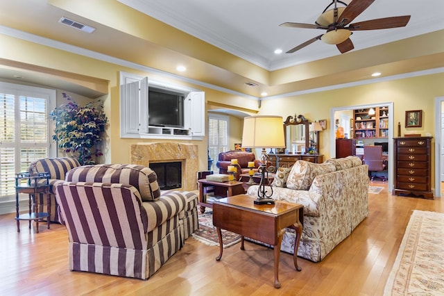 living room with crown molding, light hardwood / wood-style flooring, ceiling fan, a tray ceiling, and a premium fireplace