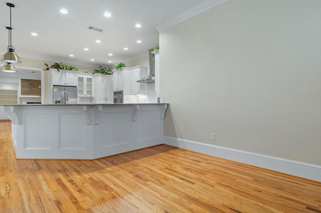 kitchen with white cabinetry, wall chimney range hood, stainless steel appliances, hanging light fixtures, and kitchen peninsula