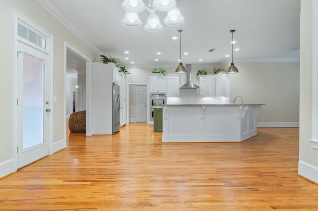 kitchen featuring hanging light fixtures, appliances with stainless steel finishes, wall chimney exhaust hood, white cabinetry, and a large island with sink