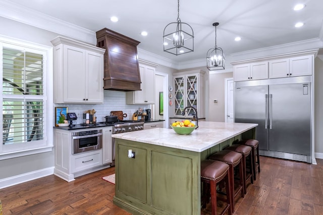 kitchen with custom exhaust hood, hanging light fixtures, a center island with sink, ornamental molding, and stainless steel appliances