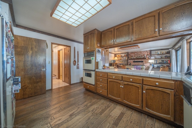 kitchen featuring dark wood-type flooring, tile counters, kitchen peninsula, brick wall, and white double oven