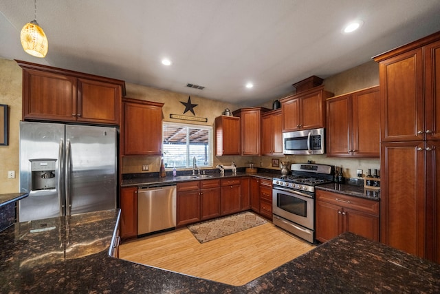 kitchen with appliances with stainless steel finishes, dark stone counters, decorative light fixtures, sink, and light hardwood / wood-style flooring