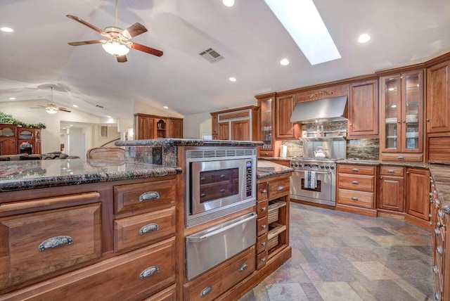 kitchen featuring vaulted ceiling with skylight, ceiling fan, backsplash, stainless steel appliances, and wall chimney exhaust hood