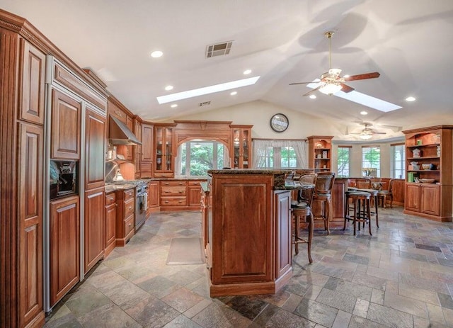 kitchen with a kitchen island, a kitchen bar, wall chimney exhaust hood, ceiling fan, and dark tile flooring