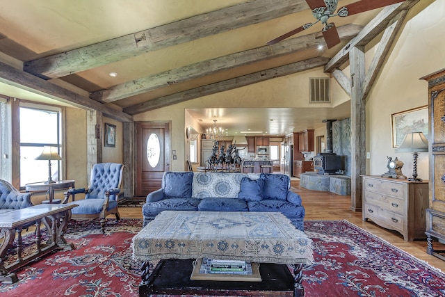 living room featuring visible vents, a wood stove, ceiling fan with notable chandelier, light wood finished floors, and vaulted ceiling with beams