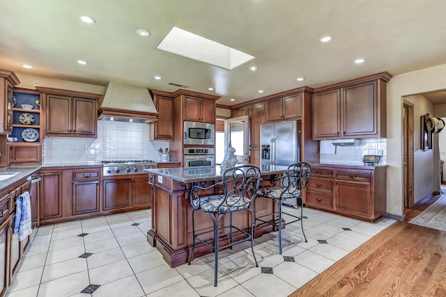 kitchen with a breakfast bar, custom exhaust hood, a skylight, dark stone counters, and stainless steel appliances