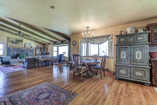 dining space with wood finished floors, baseboards, vaulted ceiling with beams, a stone fireplace, and ceiling fan with notable chandelier