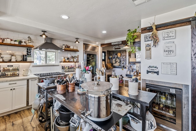 kitchen featuring light wood-type flooring, white cabinets, beverage cooler, decorative backsplash, and wall chimney range hood