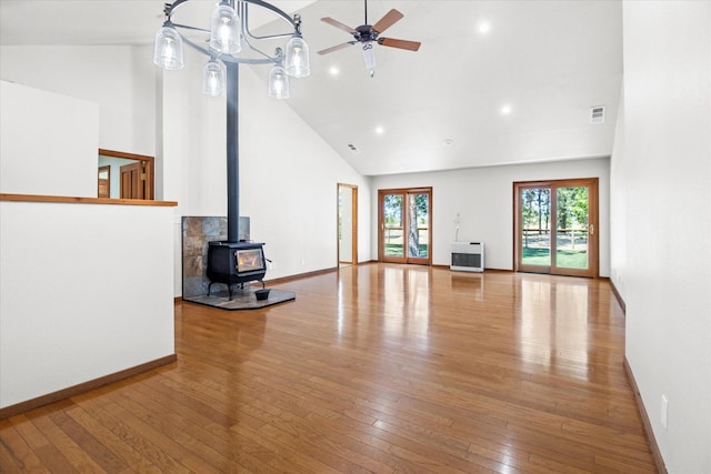 unfurnished living room featuring high vaulted ceiling, ceiling fan with notable chandelier, wood-type flooring, and a wood stove