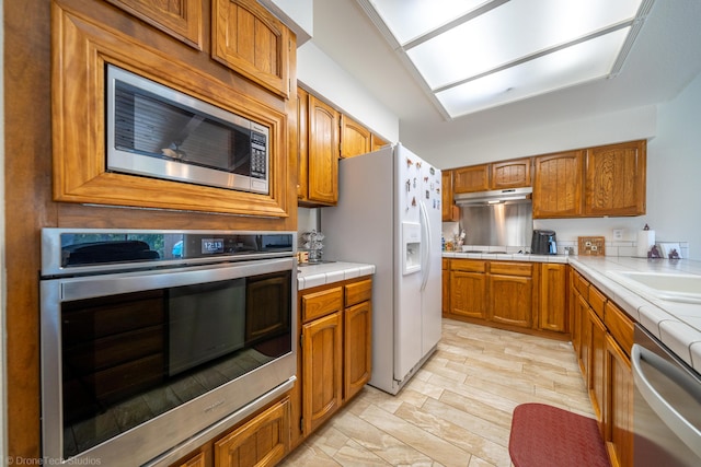 kitchen featuring appliances with stainless steel finishes, under cabinet range hood, brown cabinets, and tile countertops