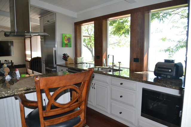 kitchen featuring wall chimney range hood, white cabinetry, sink, and dark stone countertops