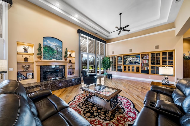 living room with ceiling fan, a raised ceiling, a high ceiling, wood-type flooring, and a premium fireplace