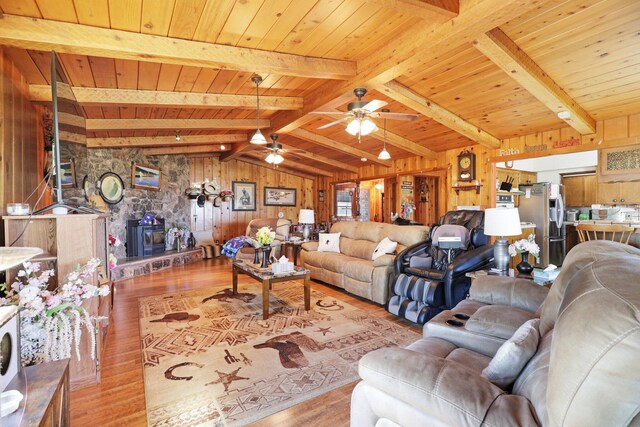 living room featuring light wood-type flooring, wood walls, ceiling fan, a stone fireplace, and lofted ceiling with beams