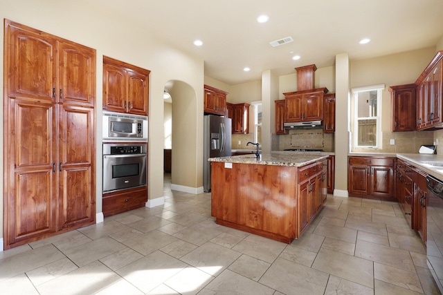 kitchen featuring appliances with stainless steel finishes, decorative backsplash, light stone countertops, a center island with sink, and sink