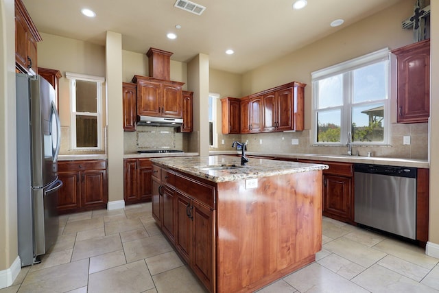 kitchen with decorative backsplash, an island with sink, light stone counters, stainless steel appliances, and sink