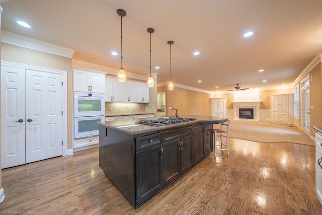 kitchen with white cabinetry, white double oven, light hardwood / wood-style flooring, stainless steel gas stovetop, and a center island with sink