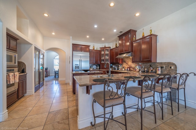 kitchen with light tile patterned flooring, built in appliances, sink, decorative backsplash, and dark stone countertops