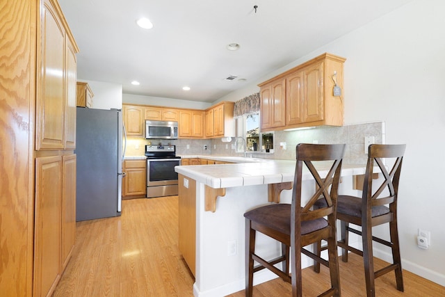 kitchen with kitchen peninsula, a kitchen breakfast bar, stainless steel appliances, tile counters, and light hardwood / wood-style floors