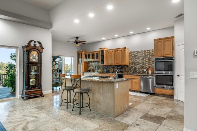 kitchen featuring backsplash, appliances with stainless steel finishes, a breakfast bar, an island with sink, and dark stone counters