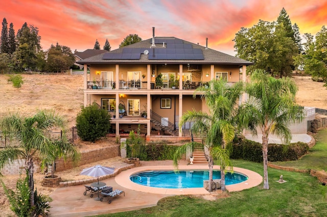 back house at dusk featuring a patio, a balcony, a fenced in pool, a lawn, and solar panels