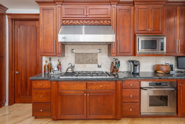 kitchen with tasteful backsplash, exhaust hood, appliances with stainless steel finishes, light hardwood / wood-style flooring, and dark stone countertops