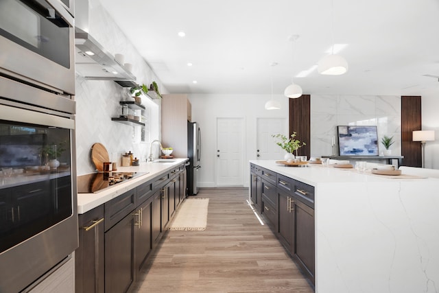 kitchen featuring wall chimney range hood, light wood-type flooring, stainless steel appliances, sink, and decorative light fixtures