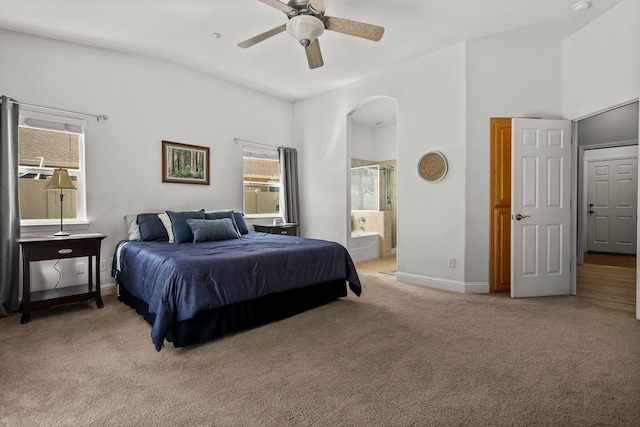 bedroom featuring arched walkways, baseboards, light colored carpet, ceiling fan, and ensuite bath