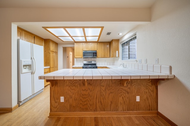 kitchen with stainless steel gas stove, white fridge with ice dispenser, kitchen peninsula, tile countertops, and light wood-type flooring