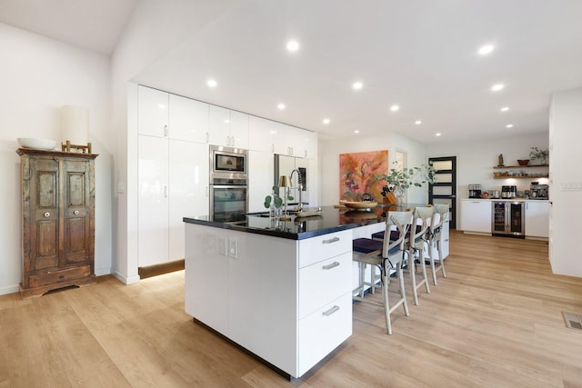 kitchen with white cabinets, an island with sink, beverage cooler, and light wood-type flooring