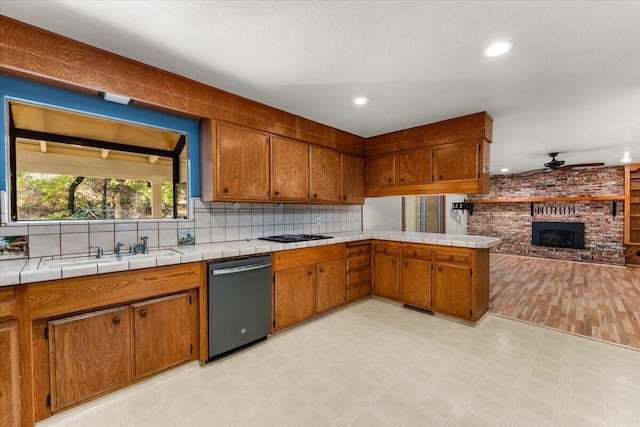 kitchen featuring ceiling fan, tile counters, dishwasher, sink, and a brick fireplace