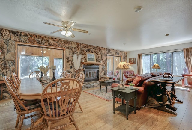 dining space featuring a textured ceiling, light hardwood / wood-style floors, a stone fireplace, and ceiling fan